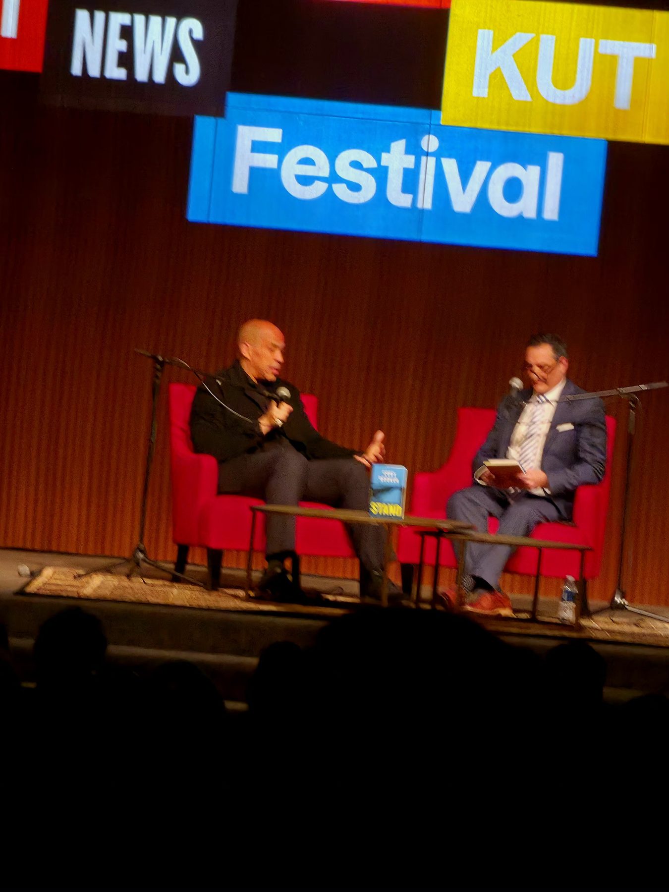 Two people seated in armchairs on a stage under warm lighting, mid conversation, with a screen behind them.