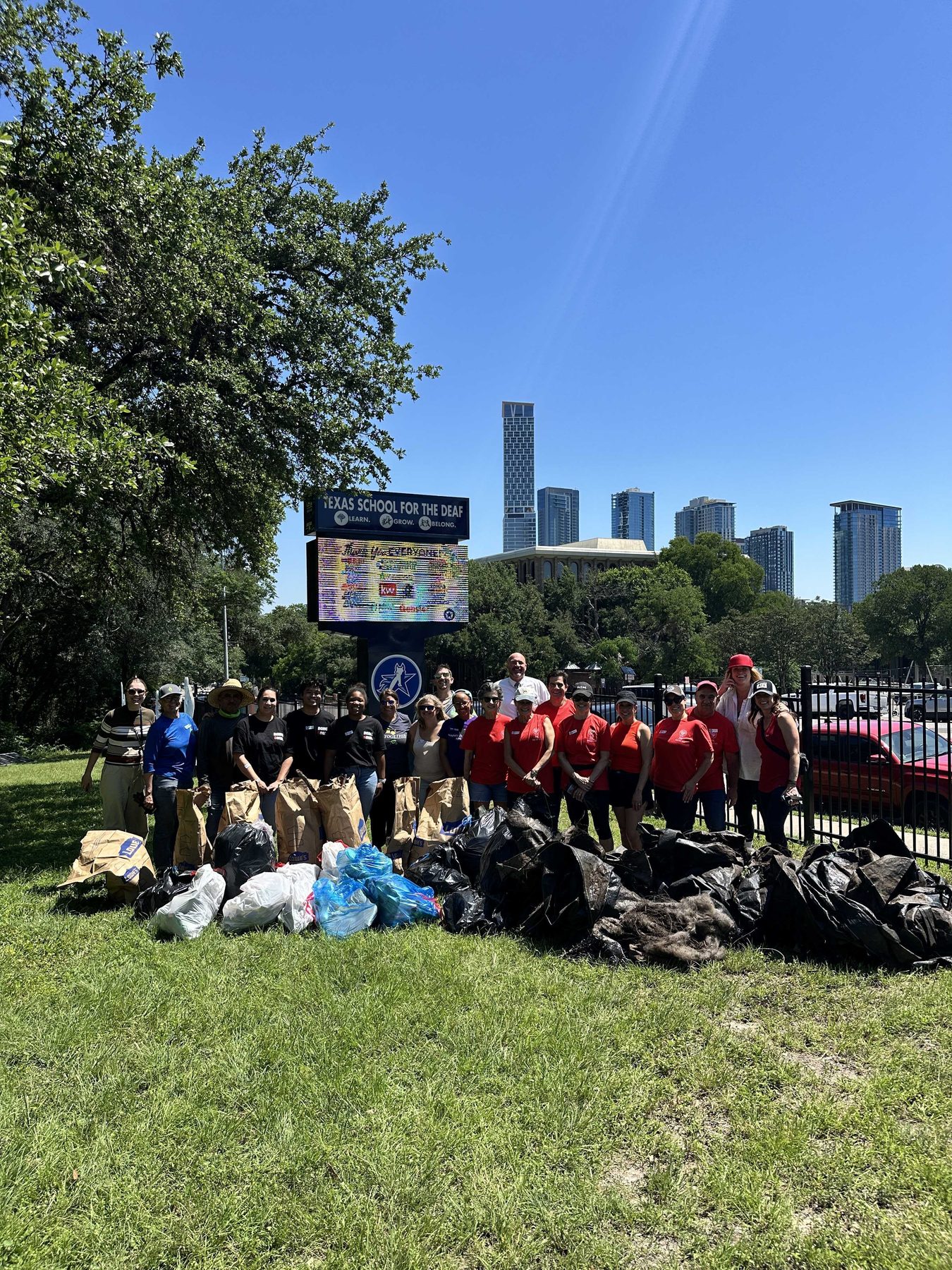 A group of volunteers standing behind a pile of filled bags on a grass verge, with downtown towers rising behind the trees.