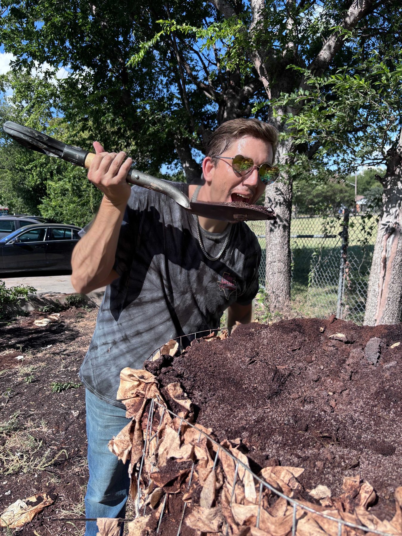 A person holding up a shovelful of dark composted coffee grounds towards their open mouth, grinning, beside a wire bin heaped with the same material and spent paper filters.