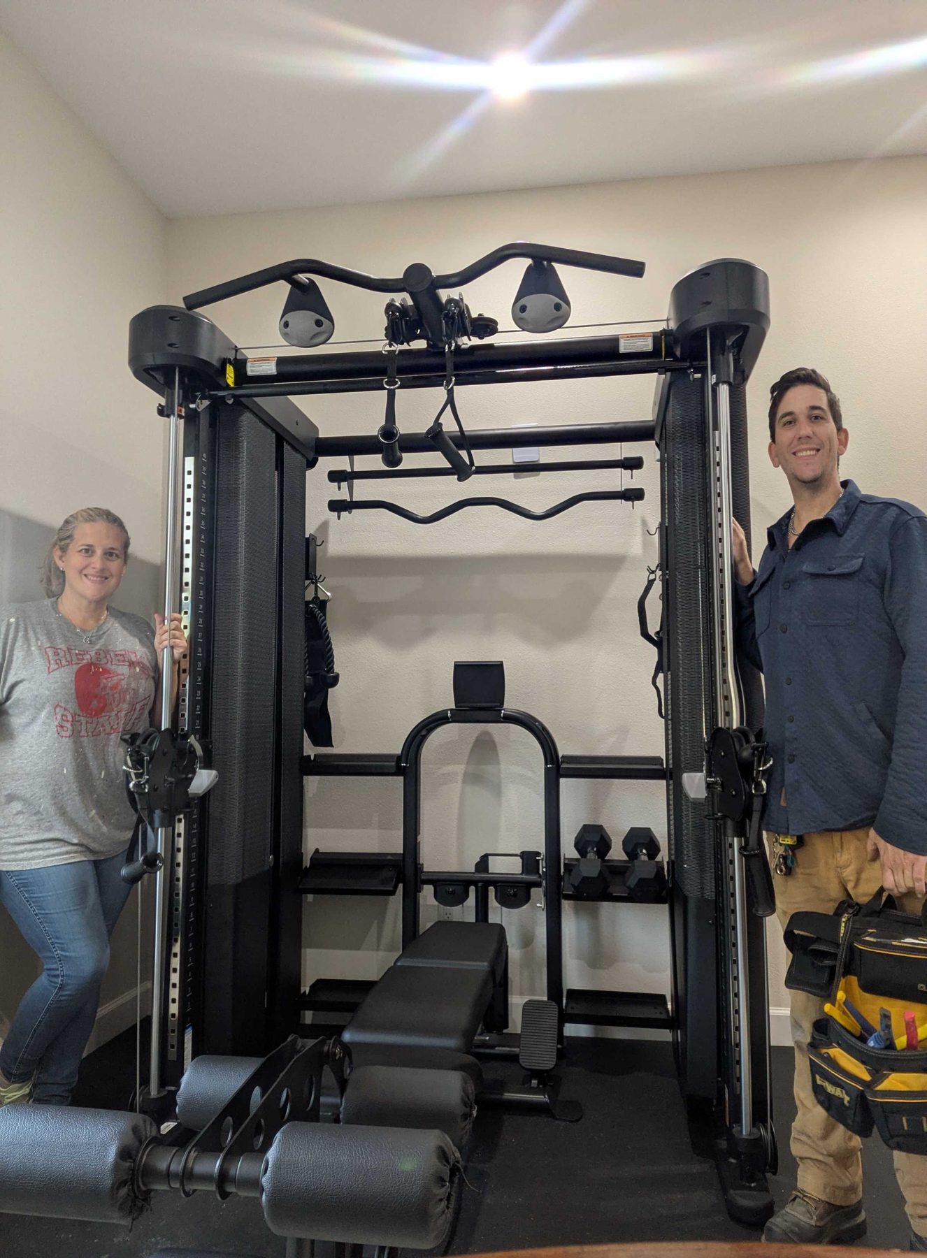 Two people standing either side of a home gym rack they have just finished assembling, with a tool bag open on the floor.