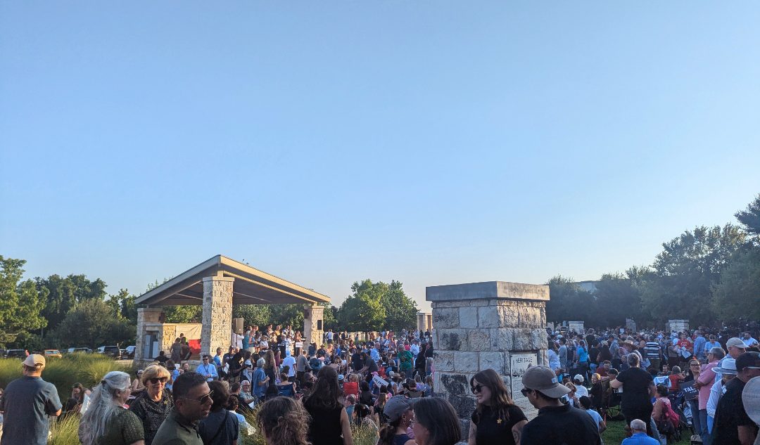 A large crowd filling a grassy field in front of an open stone pavilion under a clear sky.