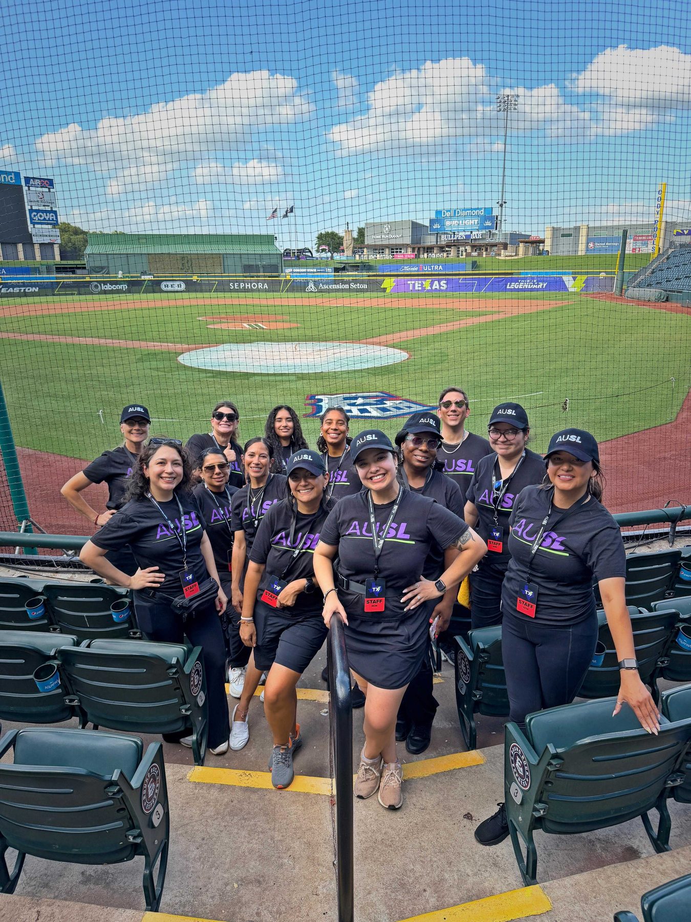 A team in matching dark shirts and caps standing together on the seating at a baseball stadium, the field and scoreboard behind them.