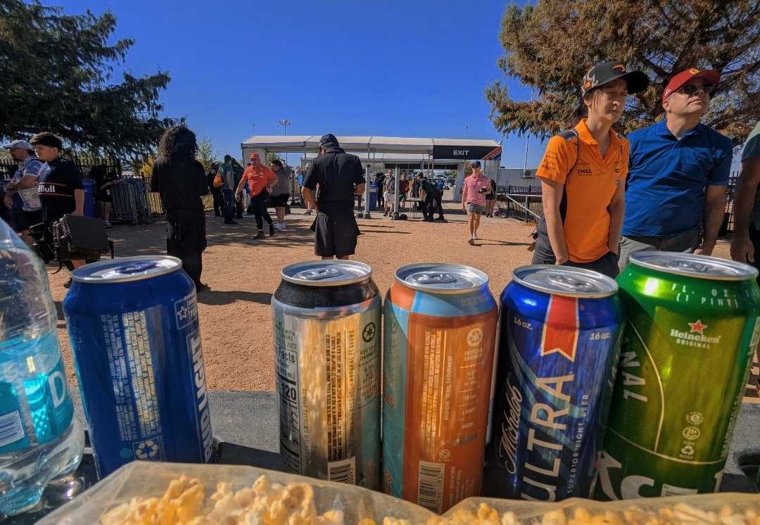 Tall drink cans lined up along the front of an outdoor service counter, with an event crowd and trees in the background.