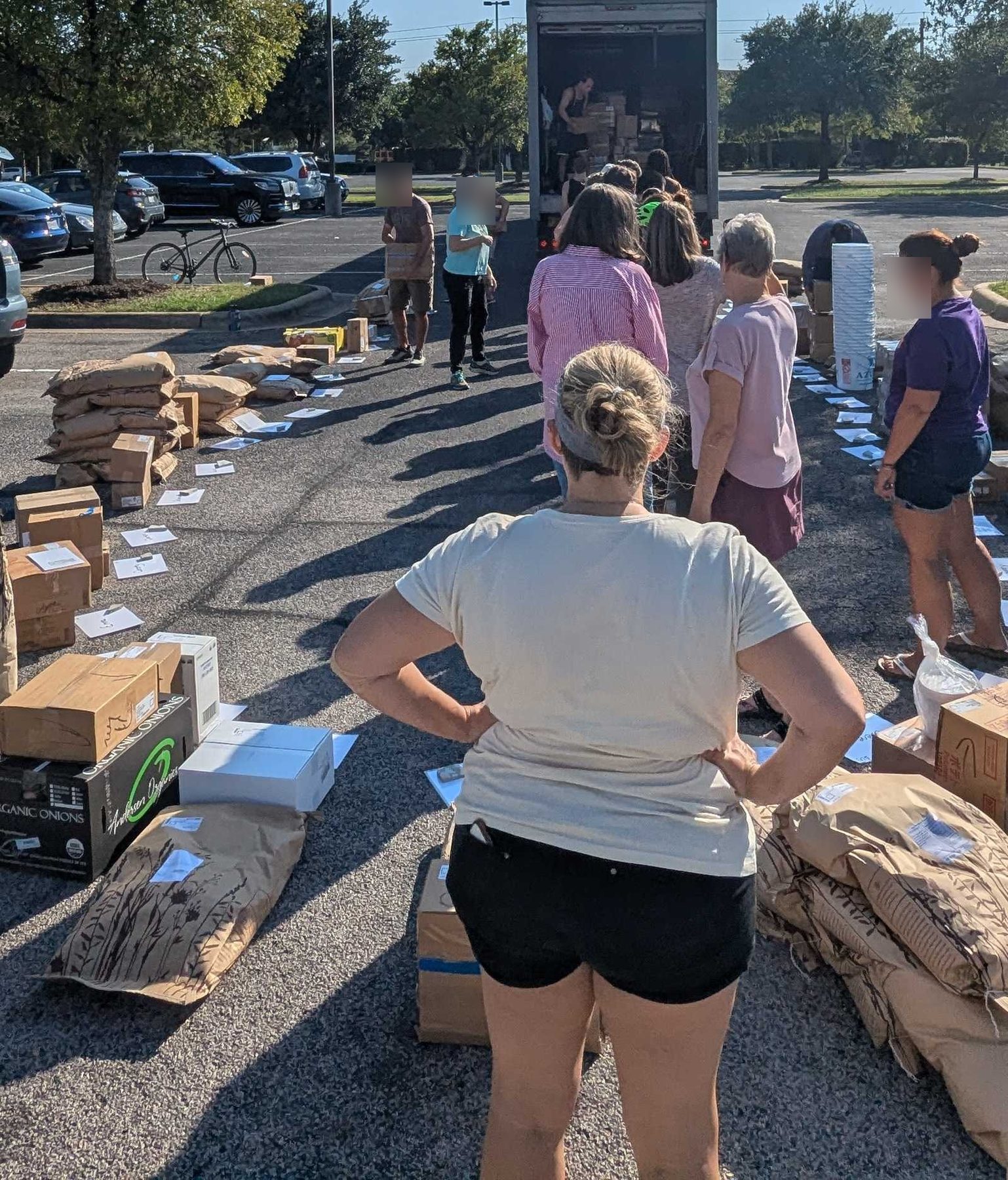 Boxes and sacks laid out in long rows across a parking lot beside an open box truck, with a line of people moving along them.