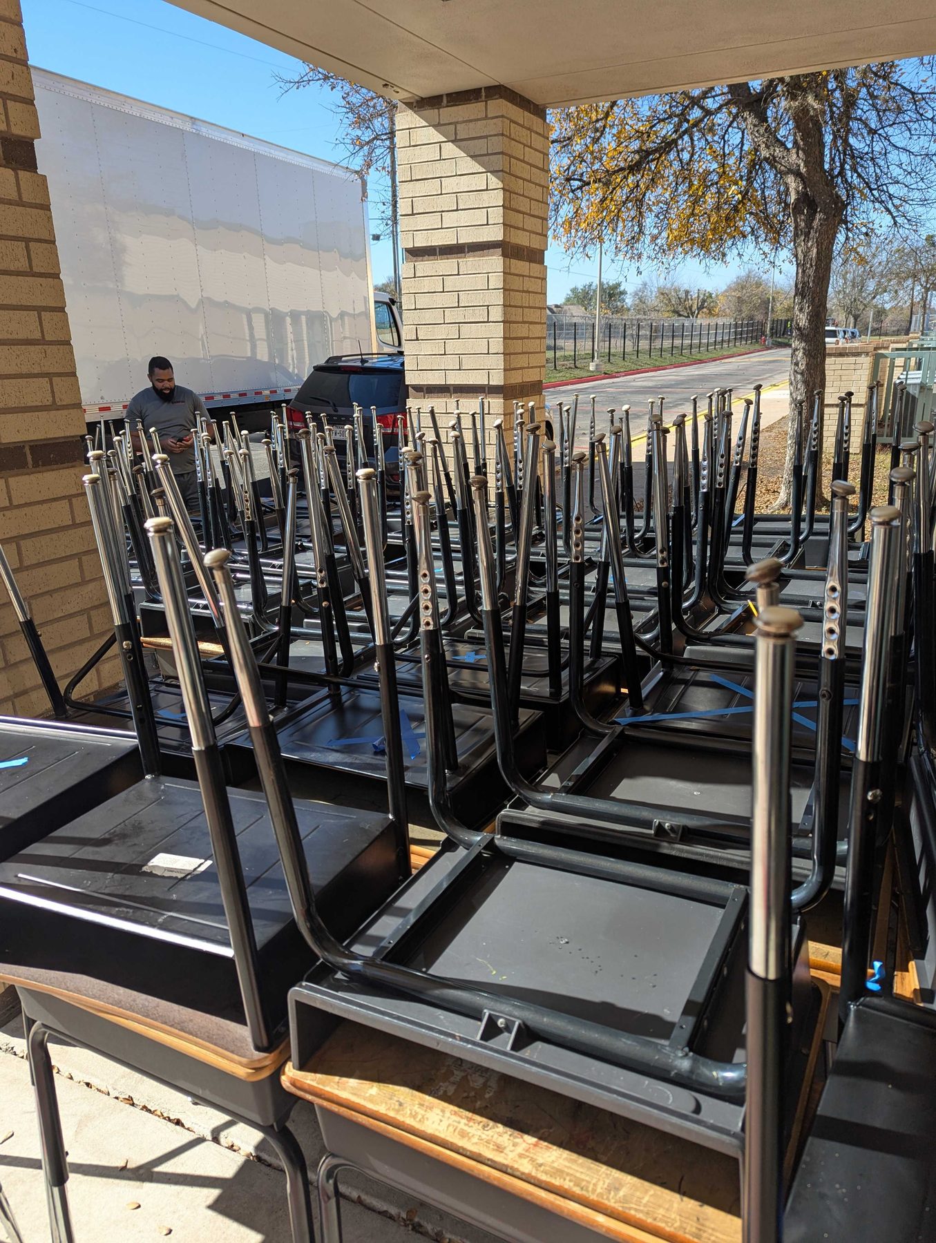 Stacked school desks and folded tables under a covered walkway, part way through being moved out, with someone working behind the pile.