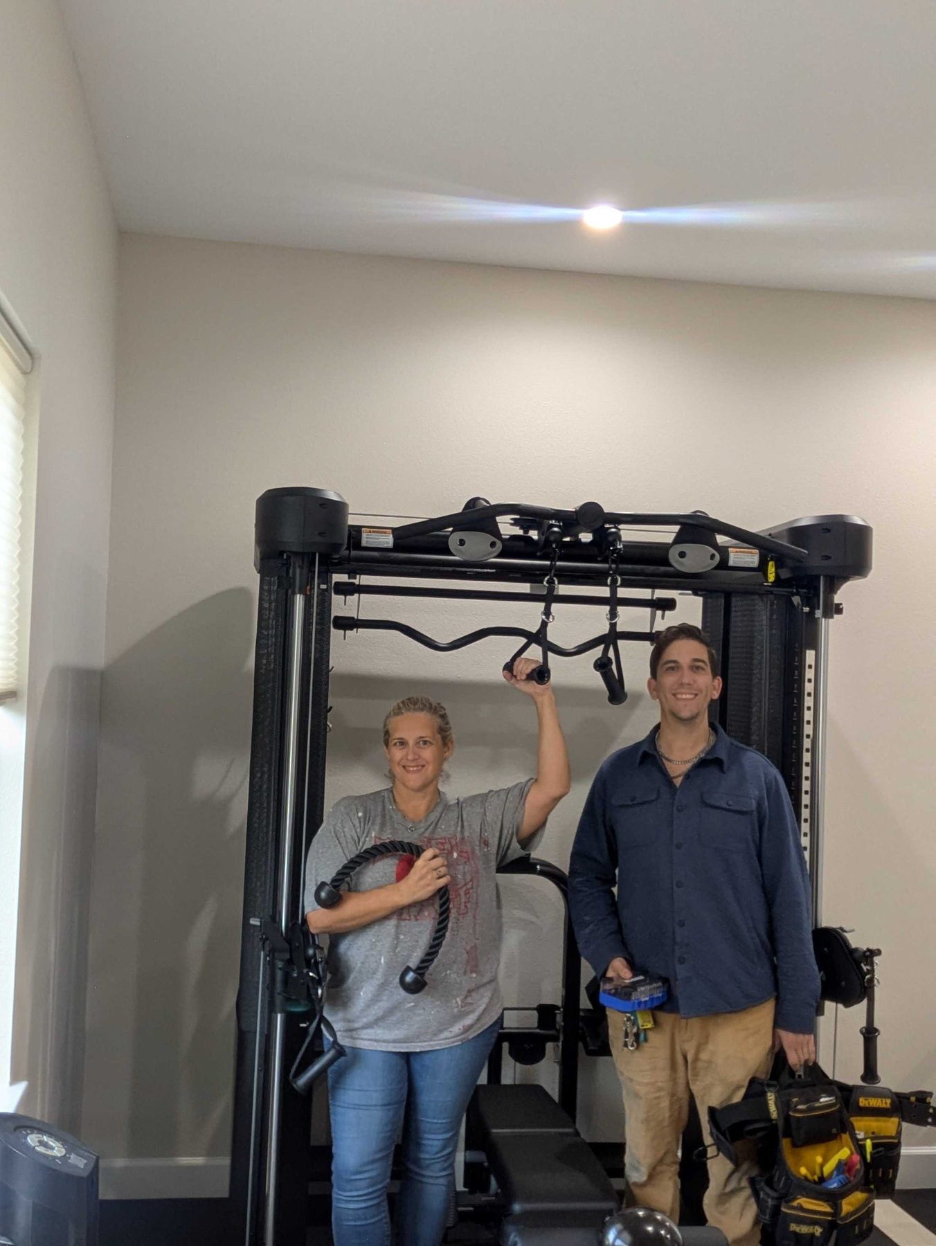 Two people beside a fully assembled home gym rack in a spare room, one holding the cable attachment, a loaded tool bag beside them.