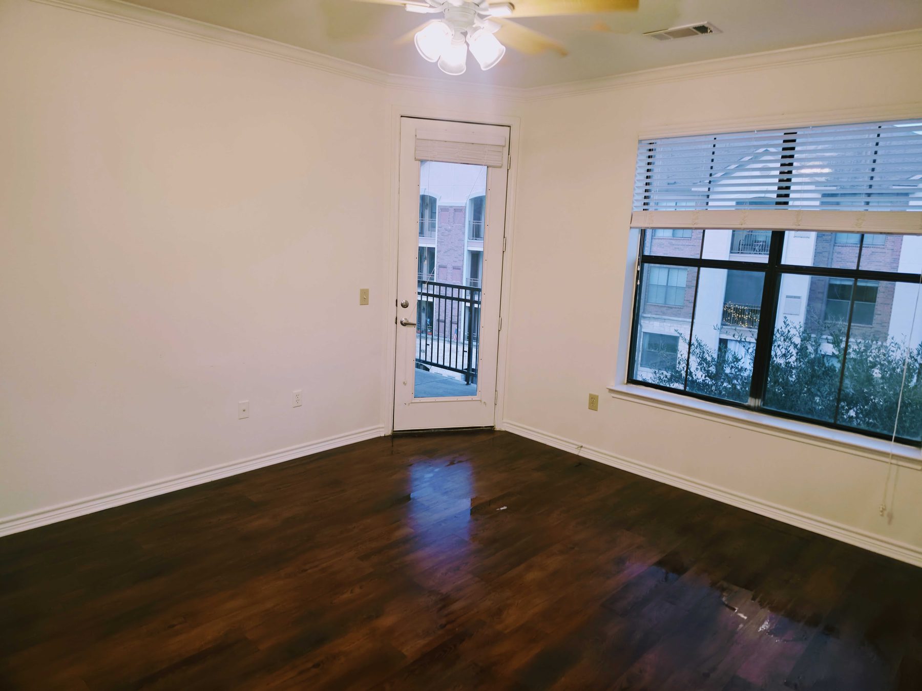 An emptied apartment room with dark wood floors, bare walls, and a balcony door standing open to the light.