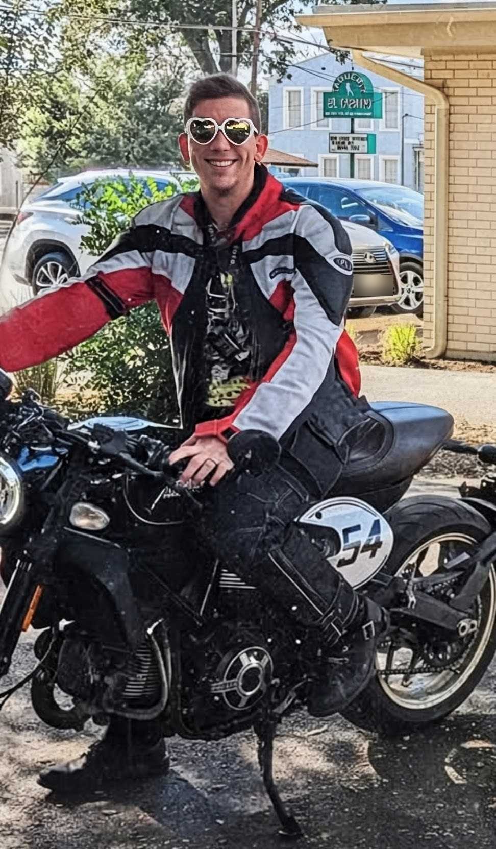 A rider in full red, gray and black race leathers sitting on a black motorcycle with a number 54 side plate, parked on a gravel lot in the sun.
