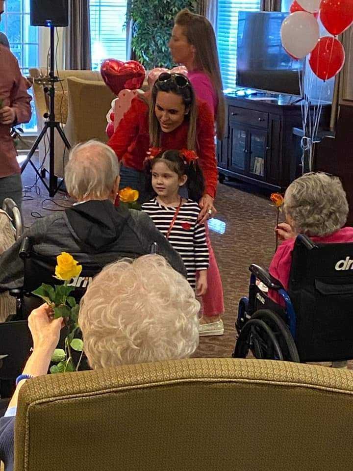A gathering at a care home, residents holding flowers and balloons while visitors sit and talk with them.
