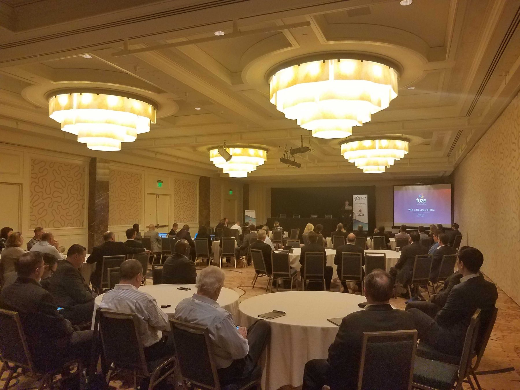 A conference room part way through a session, round tables occupied and a screen lit at the front.