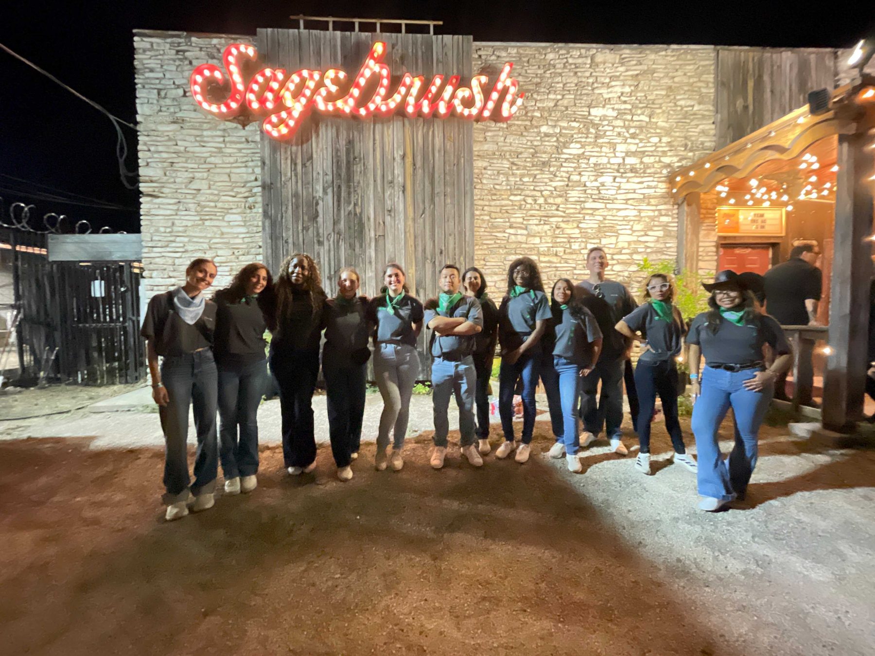 A group of people standing shoulder to shoulder outside a venue at night, a lit sign on the stone wall behind them.