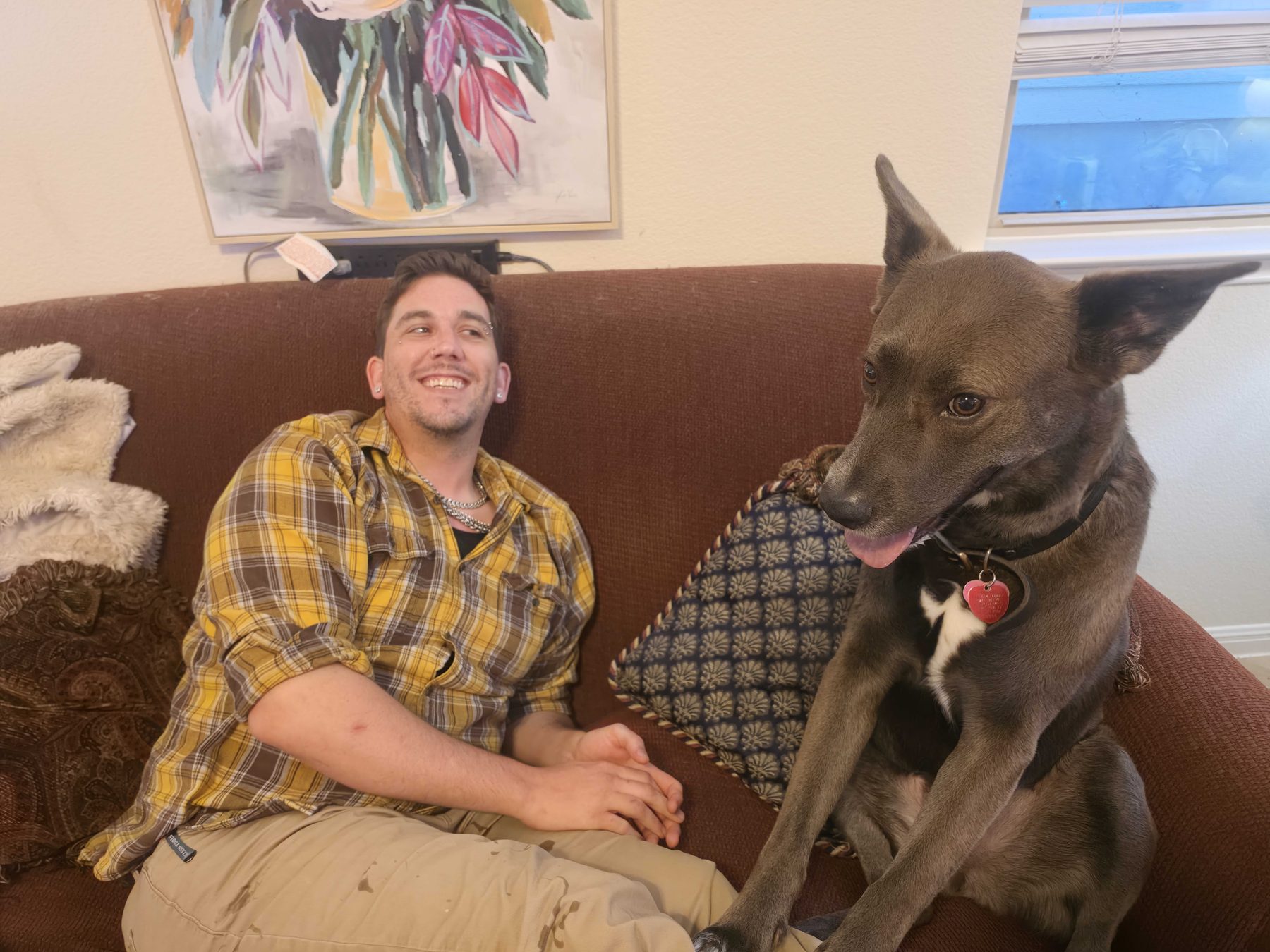 A person sitting on a couch at home beside a large gray dog, both settled and looking at the camera.